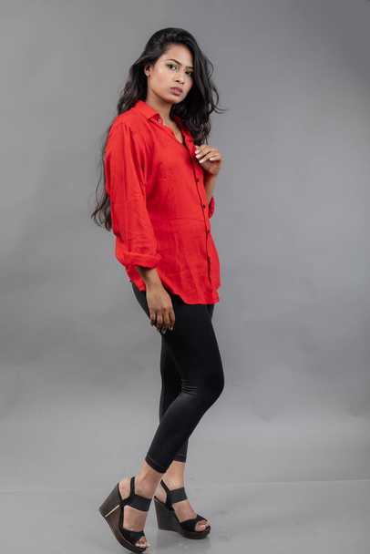 Modello giovane donna indiana in abbigliamento casual su sfondo grigio - foto stock. Modello lungo dai capelli neri con camicia rossa e jeans neri. - Foto, immagini