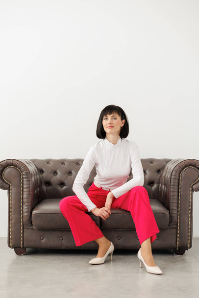 Положительная брюнетка среднего возраста женщина носит элегантные красные брюки, белую блузку и высокие каблуки уверенно смотрит в камеру, сидит спокойно на классическом кожаном диване в пустой комнате студии. Всемирный женский день. - Фото, изображение