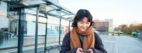 Joyeux sourire coréen fille, en utilisant un téléphone mobile, debout sur l'arrêt de bus avec smartphone, regardant l'horaire de départ sur demande, posant en vêtements d'hiver. - Photo, image