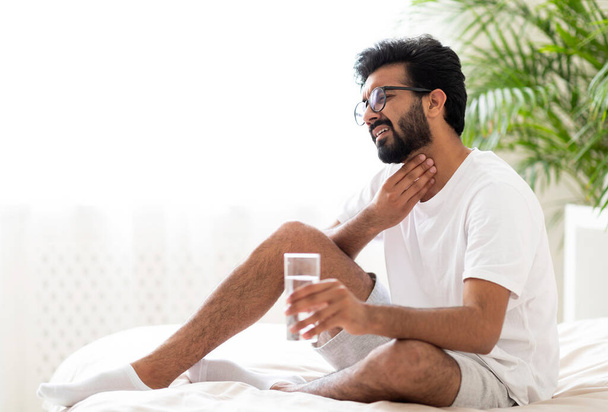 Sick young indian man suffering throat ache while sitting on bed at home, Ill eastern man touching neck with hands and frowning, having acute pain, holding glass with water, copy space - Photo, Image