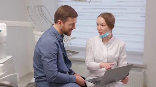 A handsome young man is talking to a female doctor at a dental appointment in a bright, beautiful office. The dentist explains to the patient and shows everything on the laptop - Footage, Video