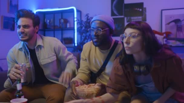Medium shot of Caucasian girl and two African American and Middle Eastern guys sitting on couch, anxiously watching sport match, cheering, whooping at scored goal, hugging and doing high fives - Footage, Video