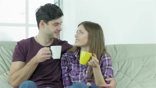 Пара дивиться один на одного, сидячи на дивані з чашками. Щасливі друзі розважаються разом. Весела молодь сидить на дивані і п'є чай або каву, розважаючись і роблячи себе - Кадри, відео