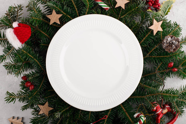 Kerst tafel instelling met witte keramische plaat op natuurlijke sparren takken achtergrond. Kerst feestelijke decoratie serveren voor Kerstmis diner, platte lay, bovenaanzicht, wenskaart template - Foto, afbeelding