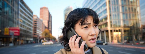 Close up portrait of concerned asian woman, talks on mobile phone and hears bad news, looks worried, feels frustrated by telephone conversation, stands on street. - Foto, Imagem