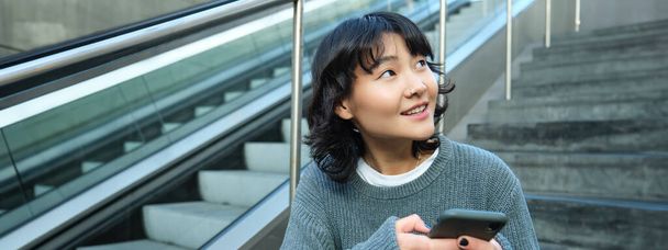 Tyylikäs nuori tyttö opiskelija, istuu portaissa älypuhelimen ja reppu, nauraa ja hymyilee, tekstiviesti, käyttää sosiaalista mediaa. - Valokuva, kuva