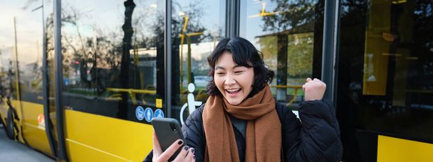 Muotokuva iloinen aasialainen tyttö puhuu matkapuhelimeen, video chatit, näyttää hämmästynyt älypuhelin kamera, seisoo bussipysäkillä. - Valokuva, kuva