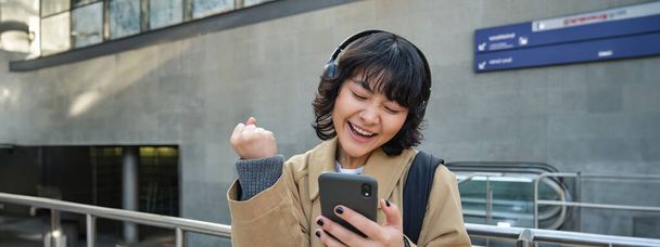 Cheerful Korean girl wins, celebrates victory, looks at smartphone app and triumphs, express joy and excitement, stands in headphones on empty street. - Zdjęcie, obraz