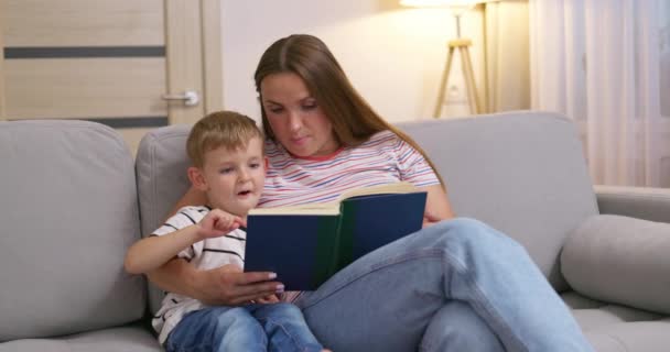 Mère et son fils lisent un livre assis sur un canapé confortable dans le salon, du temps en famille. Images 4k de haute qualité - Séquence, vidéo