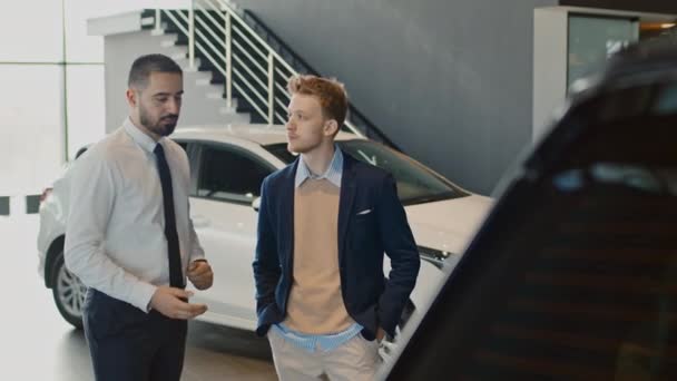 Medium full shot of Middle Eastern salesman in white shirt and tie talking to Caucasian businessman in smart casual clothes in car dealership and explaining features of new crossover model - Footage, Video