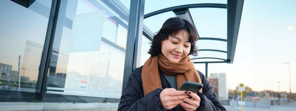 Belle fille japonaise, étudiante debout sur l'arrêt de bus en veste d'hiver, tenant smartphone, en utilisant l'application pour suivre ses transports en commun, commander un tour. - Photo, image