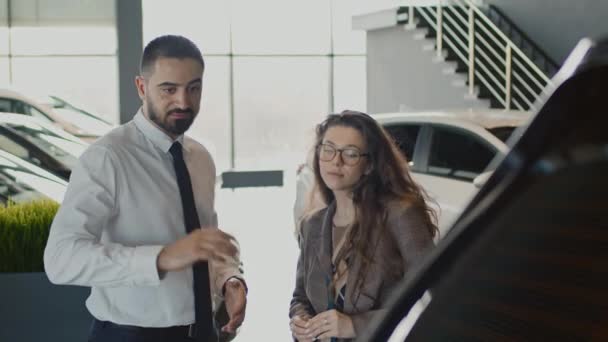 Średnie ujęcie młodej białej kobiety wybierającej nowy samochód w salonie samochodowym i słuchającej męskiego przedstawiciela handlowego, który wyjaśnia charakterystykę nowej modyfikacji zwrotnicy - Materiał filmowy, wideo