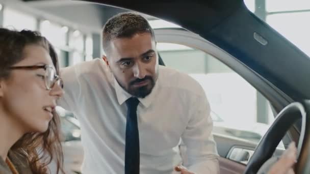 Medium close-up shot of happy young Caucasian woman sitting in driver seat of new car in automobile dealership, touching ergonomic steering wheel, and salesman explaining operational features - Footage, Video