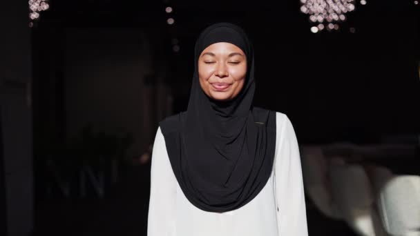 Vista frontale di una bella donna afroamericana vestita in hijab nero che posa in un luminoso ufficio moderno con braccia incrociate. Donna d'affari di successo con un sorriso sincero sul viso guardando la fotocamera sul posto di lavoro. - Filmati, video