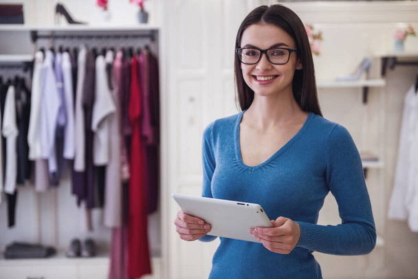 Linda assistente de loja feminina em óculos está usando um tablet digital, olhando para a câmera e sorrindo enquanto está em pé na boutique
 - Foto, Imagem