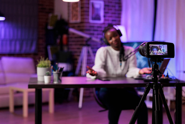 Focus shot on modern camera recording vlogger in blurry background talking about parenting subjects. Woman filming online show using professional gadgets in rgb lights studio - Photo, Image
