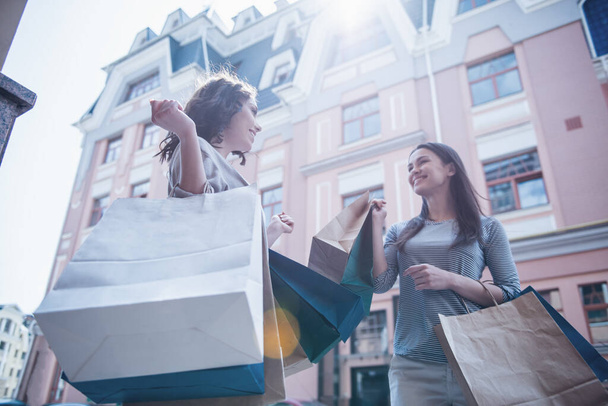 Visão de baixo ângulo de meninas bonitas segurando sacos de compras, falando e sorrindo enquanto estão de pé ao ar livre
 - Foto, Imagem