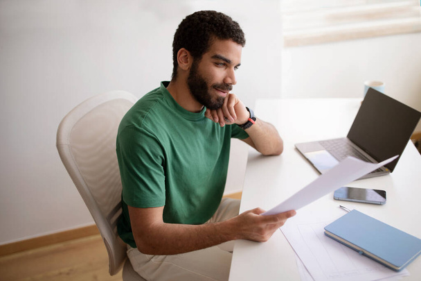 Sonriente joven islámico guapo pensativo en camiseta verde leer documentos, trabajar con el ordenador portátil con pantalla vacía, en el interior de la oficina en casa. Contrato, puesta en marcha, negocio con dispositivo - Foto, imagen