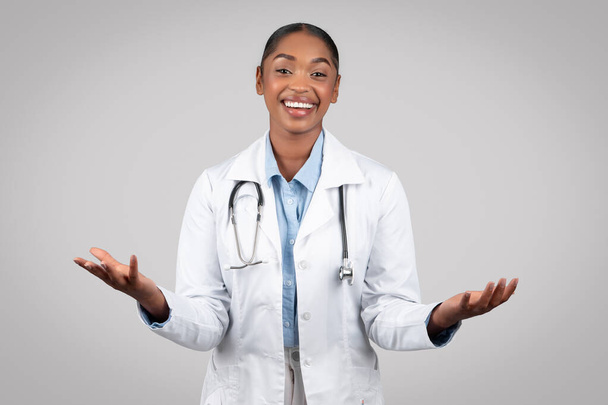 笑顔の大人のアフリカ系アメリカ人女性医師は,グレーの背景,スタジオで孤立した手の空きスペースを保持しています. 医療支援,現代医療の推奨事項,選択,広告,提供 - 写真・画像