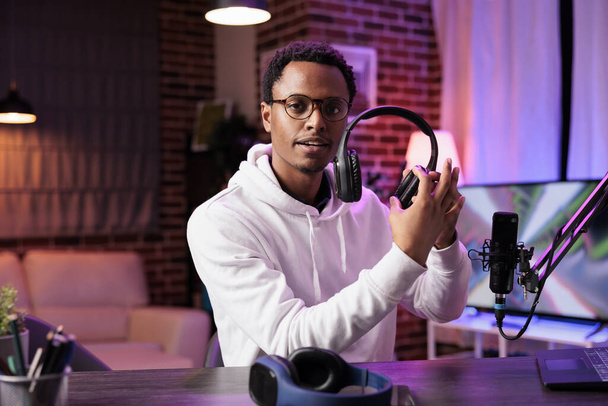 Retrato de vlogger de tecnologia americana africana revisando fones de ouvido no estúdio da sala de estar. Entretenimento criador de conteúdo filmando vlog canal de tecnologia para audiófilos, apresentando informações - Foto, Imagem