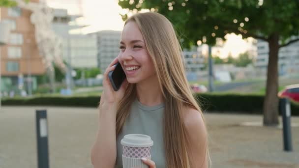 Jovencita alegre con taza de café en la mano ríe hablando por teléfono con su novio. Lady pasa el tiempo bebiendo café en el parque de la ciudad después del trabajo - Imágenes, Vídeo