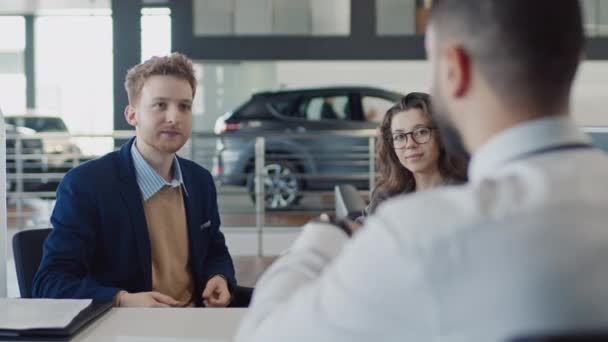 Medium over-shoulder shot of happy young man giving back signed contract to sales manager at car dealership, receiving electronic key, talking, and woman listening, nodding and smiling - Footage, Video