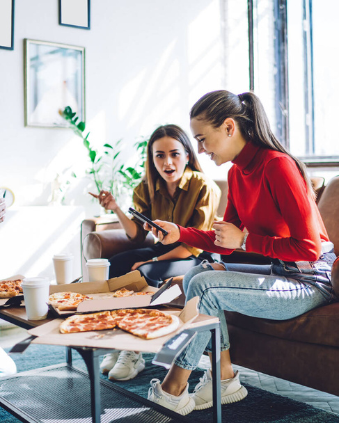 Cheerful woman in casual outfit browsing smartphone while smiling people eating delivered pizza and sitting on couch by table in stylish apartment - Photo, Image