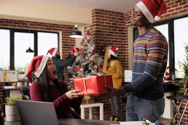 Excited asian woman employee in santa hat taking xmas gift from african american man coworker while working on laptop. Diverse colleagues exchanging presents in office at christmas eve - Photo, Image