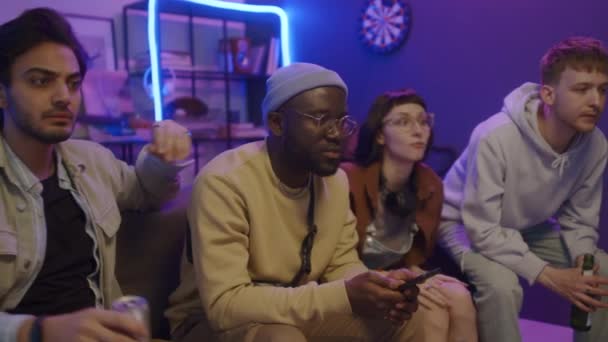 Середній знімок мультикультурних молодих студентів чоловічої та жіночої статі, що сидять у вітальні в гуртожитку, тривожно дивиться спортивний матч по телевізору, веселить, а потім реагує з розчаруванням на пропущену мету - Кадри, відео