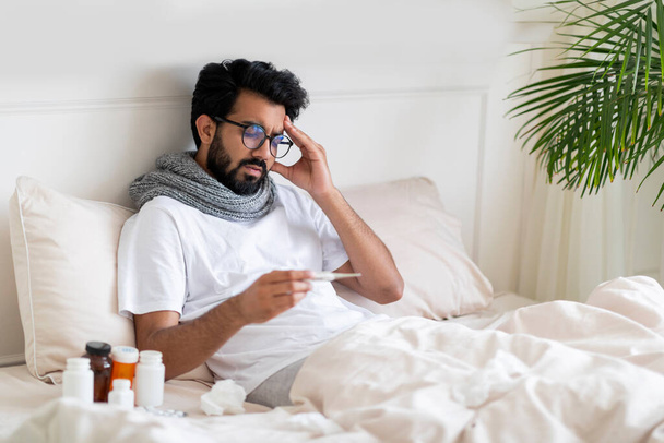 Hasta Hintli genç adam evde yatakta otururken termometreye bakıyor. Doğulu adam vücut ısısını ölçüyor ve baş ağrısına dokunuyor. Mevsimsel grip belirtileri gösteriyor. - Fotoğraf, Görsel