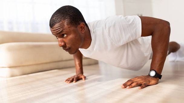 Motivado atlético jovem negro vestindo sportswear branco exercitando em sua casa ginásio, fazendo flexões, vista lateral, panorama com espaço livre. Desporto em casa - Foto, Imagem