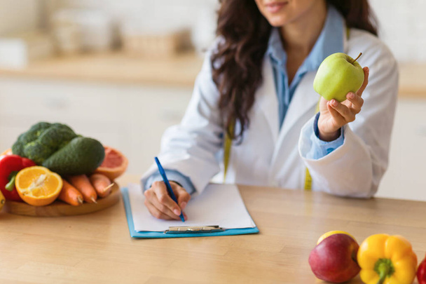 Nutricionista enfocado en pelaje blanco sentado en la mesa lleno de productos frescos, muestra manzana verde mientras observa las recomendaciones dietéticas en el portapapeles en el interior - Foto, imagen