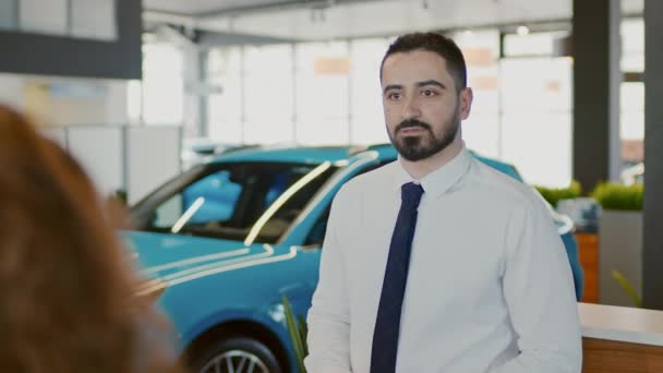 Střední přes rameno snímek středovýchodního obchodního manažera, který stojí v prodejně aut před luxusním modrým crossoverem, mluví se zákaznickým párem o svých požadavcích a odvádí je pryč - Záběry, video