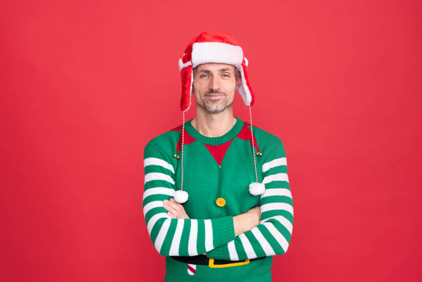 Facet w kapeluszu Świętego Mikołaja na czerwonym tle. Szczęśliwego Nowego Roku. Wesołych Świąt. szczęśliwy uśmiechnięty mężczyzna w kostiumie elfa. - Zdjęcie, obraz