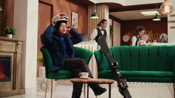 Asian girl preparing for winter sport and sitting in reception area at exclusive ski resort, checking skis and safety helmet. Winter sport enthusiast getting gear ready for skiing on snowy slopes. - Footage, Video
