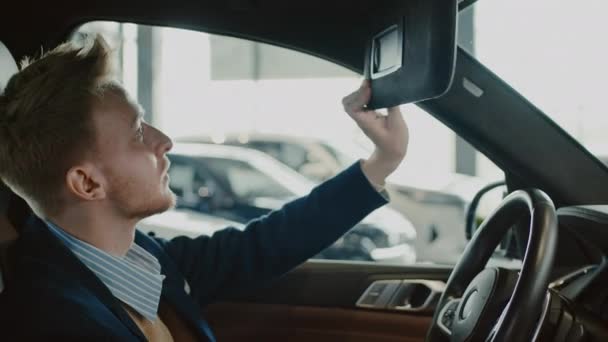 Tiro medio de joven caucásico comprador masculino sentado en el asiento del conductor del coche nuevo en la concesionaria, tocando el volante, ajustando su pelo rojo en el espejo en la visera del parabrisas, abriendo el compartimiento de almacenamiento - Imágenes, Vídeo