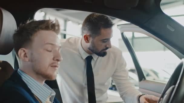Mediano primer plano de cliente masculino caucásico sentado en el asiento del conductor del coche nuevo en la concesionaria, sosteniendo el volante, gerente de ventas biracial en camisa blanca y corbata que demuestra las características del vehículo - Imágenes, Vídeo