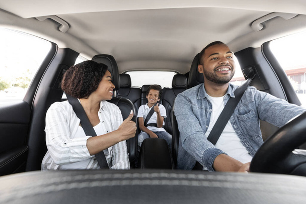 Sichere Fahrt. Glückliche schwarze Familie junge Eltern und Sohn genießen Autofahrt zusammen. Afroamerikanische Mutter schaut ihr Kind mit angeschnalltem Sicherheitsgurt an, zeigt Daumen hoch, Vater fährt Auto und lächelt - Foto, Bild