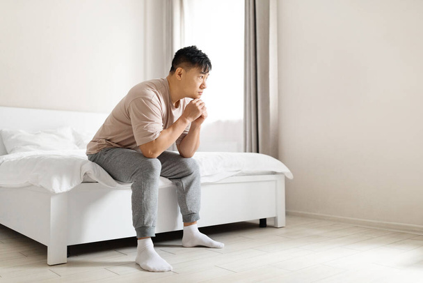 Δυσαρεστημένος απογοητευμένος ώριμος Κορεάτης που φοράει πυτζάμα στο κρεβάτι του το πρωί, ακουμπισμένος στα γόνατά του, να υποφέρει από διανοητική κατάρρευση, στο εσωτερικό του υπνοδωματίου, κοιτάζοντας το χώρο των αντιγράφων - Φωτογραφία, εικόνα