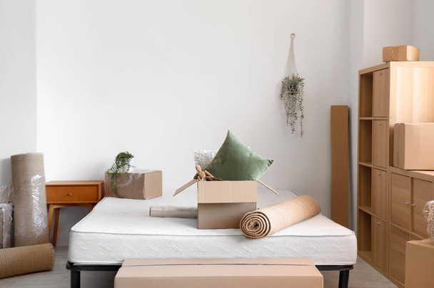 Cama con caja de cartón y alfombra enrollada en la habitación en el día en movimiento - Foto, imagen