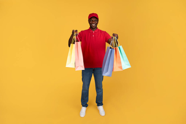 Happy millennial black guy courier hold many boxes with purchases, isolated on orange studio background. Home delivery, work and online shopping service, customer order for shopaholic - Photo, Image