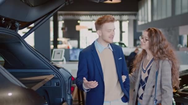 Medium shot van vrolijke jonge blanke man en vrouw onderzoeken nieuwe dure crossover in de auto showroom, man uitleggen van de kenmerken, druk op de knop en het openen van de kofferbak, en vrouw glimlachen - Video