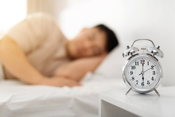 早起きするコンセプト. 朝,アジア人男性が目覚まし時計を通して眠り,ベッドに横たわり,警報時計に選択的な焦点を当てています. 目を覚まして起き上がる時間. シャロウ深度 - 写真・画像