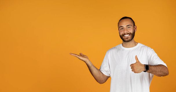 Glad millennial european man in white t-shirt show thumb up, hold on hand free space, recommends ad and offer, isolated on orange studio background. Approval gesture, positive emotions - Photo, Image