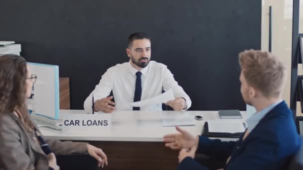 Střední záběr mužského biracial finance specialista na bílou košili a kravatu sedí za stolem v autosalonu, dává kopie smlouvy o půjčce a pojištění pro čtení mladého bělocha manžela a manželky - Záběry, video
