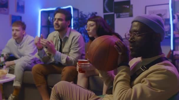 Medium shot of Caucasian girl and guy and Middle Eastern male friend sitting on couch at home, watching sport game on TV, pointing at screen and cheering, and black man fiddling with basketball - Footage, Video