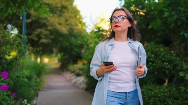Mujer feliz con laptop y smartphone en sus manos caminando por el floreciente jardín o calle verde después del día de trabajo. - Metraje, vídeo