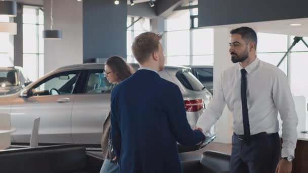 Full shot of polite Middle Eastern car salesman welcoming young Caucasian couple into showroom, inviting to sit on couch, shaking hands with husband, and giving them copies of loan agreement to read - Footage, Video