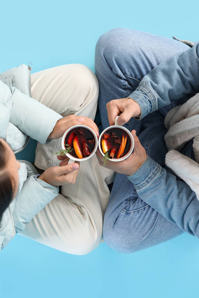Paar in warmer Kleidung mit Bechern mit leckerem Glühwein auf blauem Hintergrund sitzend, Draufsicht - Foto, Bild