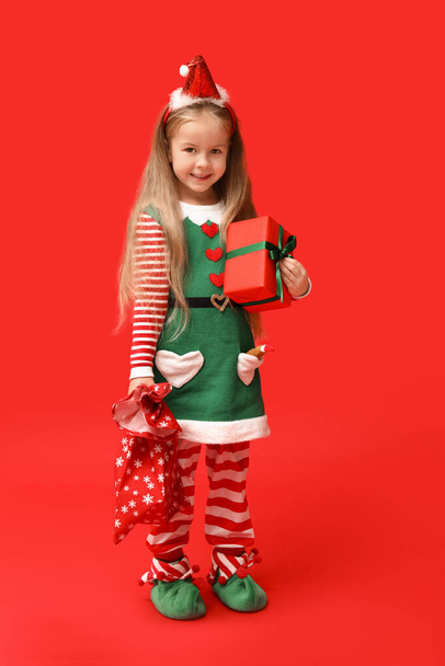 Schattig klein meisje in kerst elf kostuum met geschenkdoos en tas op rode achtergrond - Foto, afbeelding